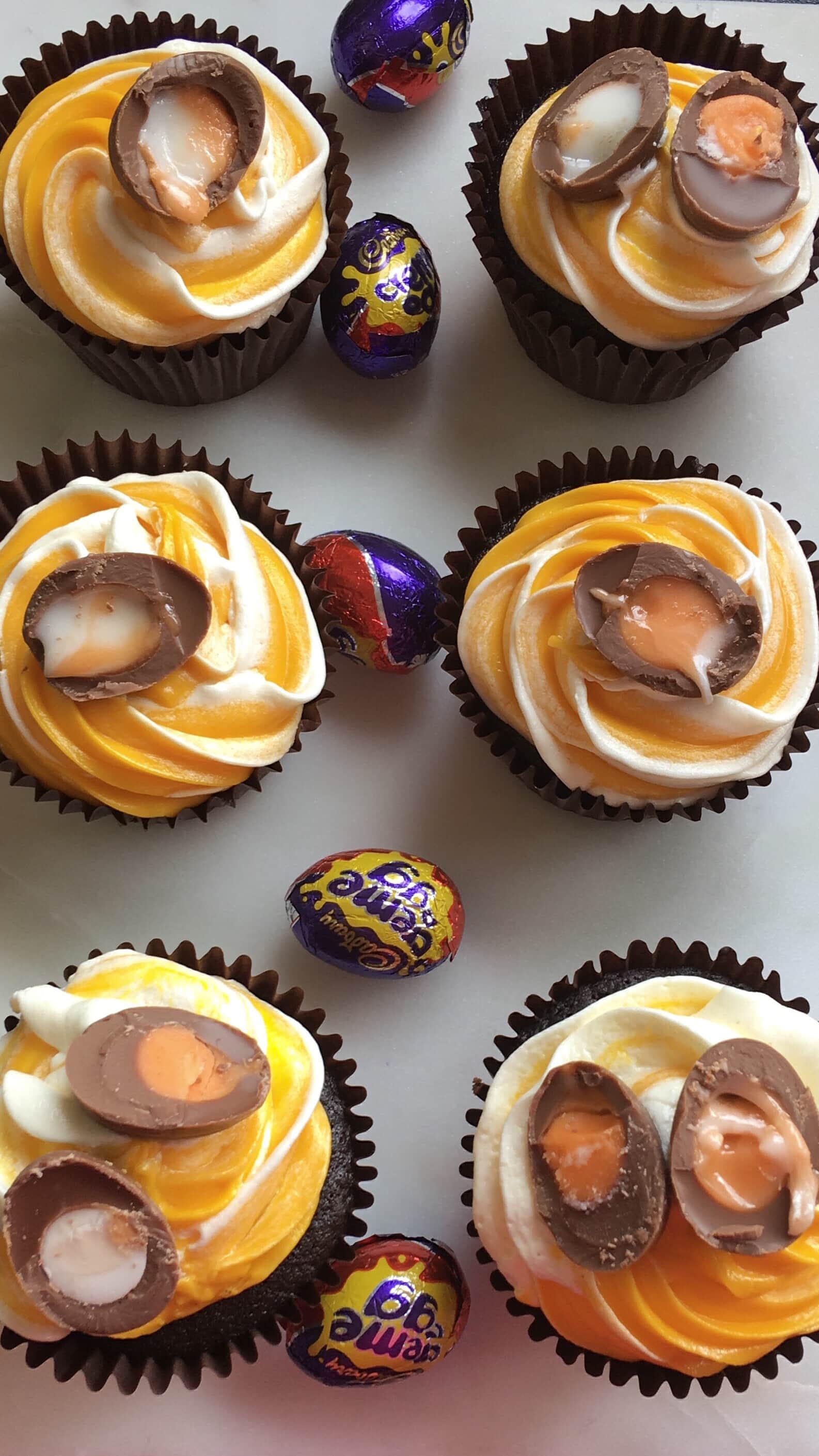 A flat lay photo of 6 chocolate cupcakes topped with yellow and white frosting and a creme egg.