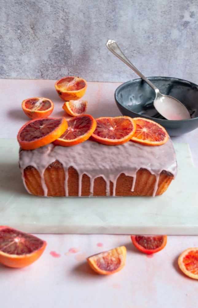 An orange loaf cake drizzled with an orange icing glaze and fresh blood orange slices on a white marbled board.  A black bowl of blood orange icing with a silver spoon and orange slices are in the background.