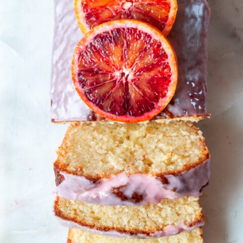a close up photo of a sliced orange loaf cake drizzled with an orange icing glaze. Fresh blood oranges are placed on top of the cake.