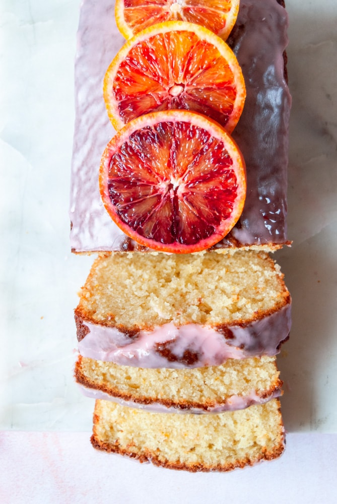 a close up photo of a sliced orange loaf cake drizzled with an orange icing glaze.  Fresh blood oranges are placed on top of the cake.