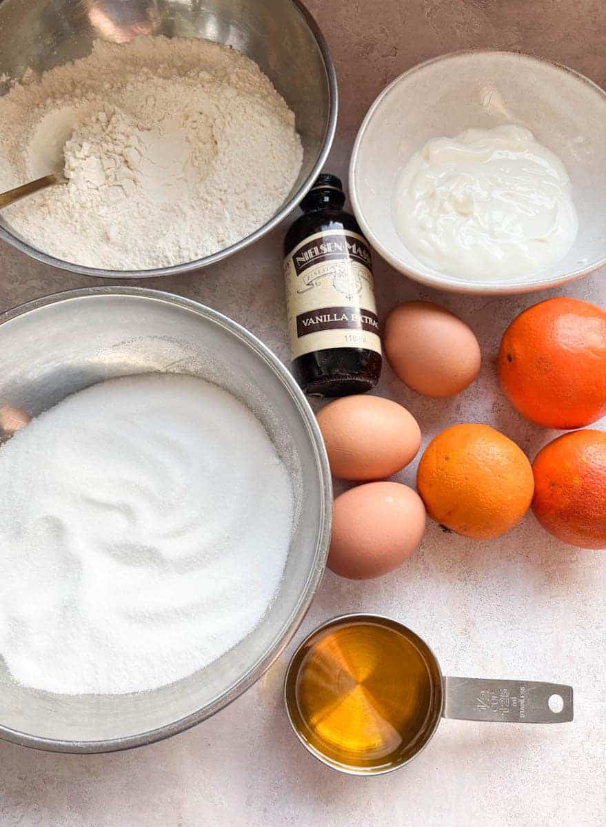 Bowls of flour, caster sugar, plain yoghurt, 3 eggs, 3 oranges, a measuring cup of olive oil and a bottle of vanilla extract.