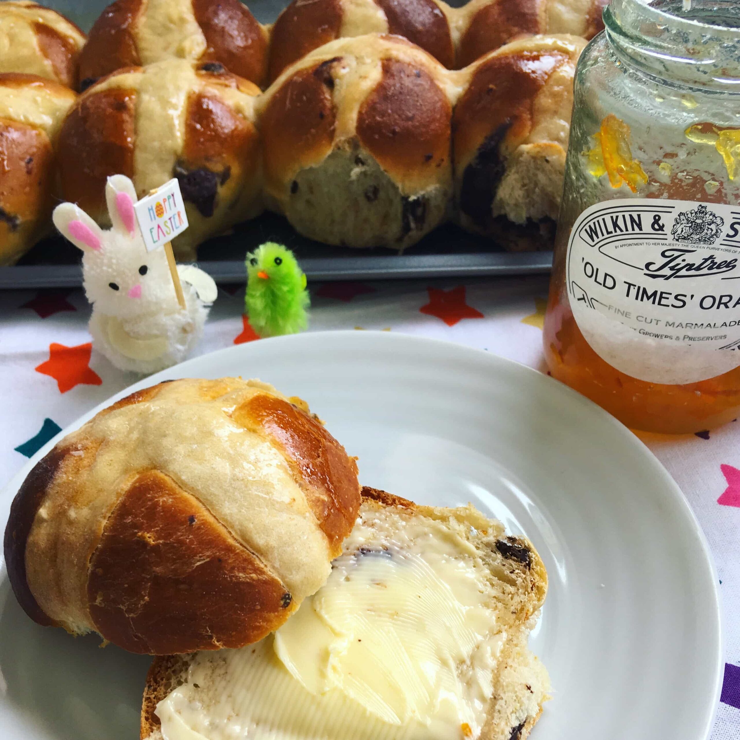 A chocolate chip hot cross bun split in half and spread with butter. A tray of hot cross buns can be seen in the background along with a jar of marmalade and a little Easter bunny and chick.