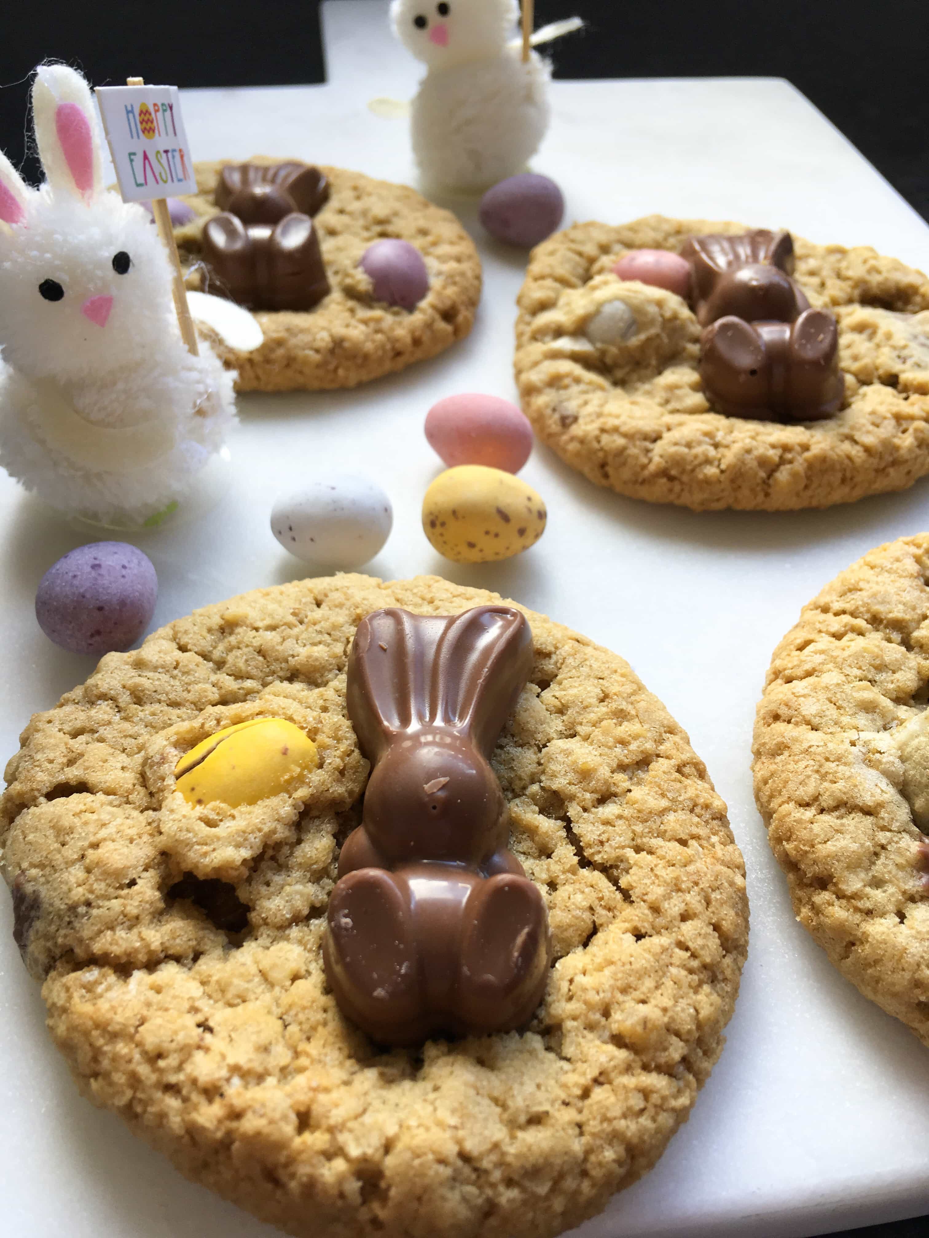 cookies topped with mini chocolate eggs and chocolate bunnies on a white chopping board with decorative Easter bunny toys.