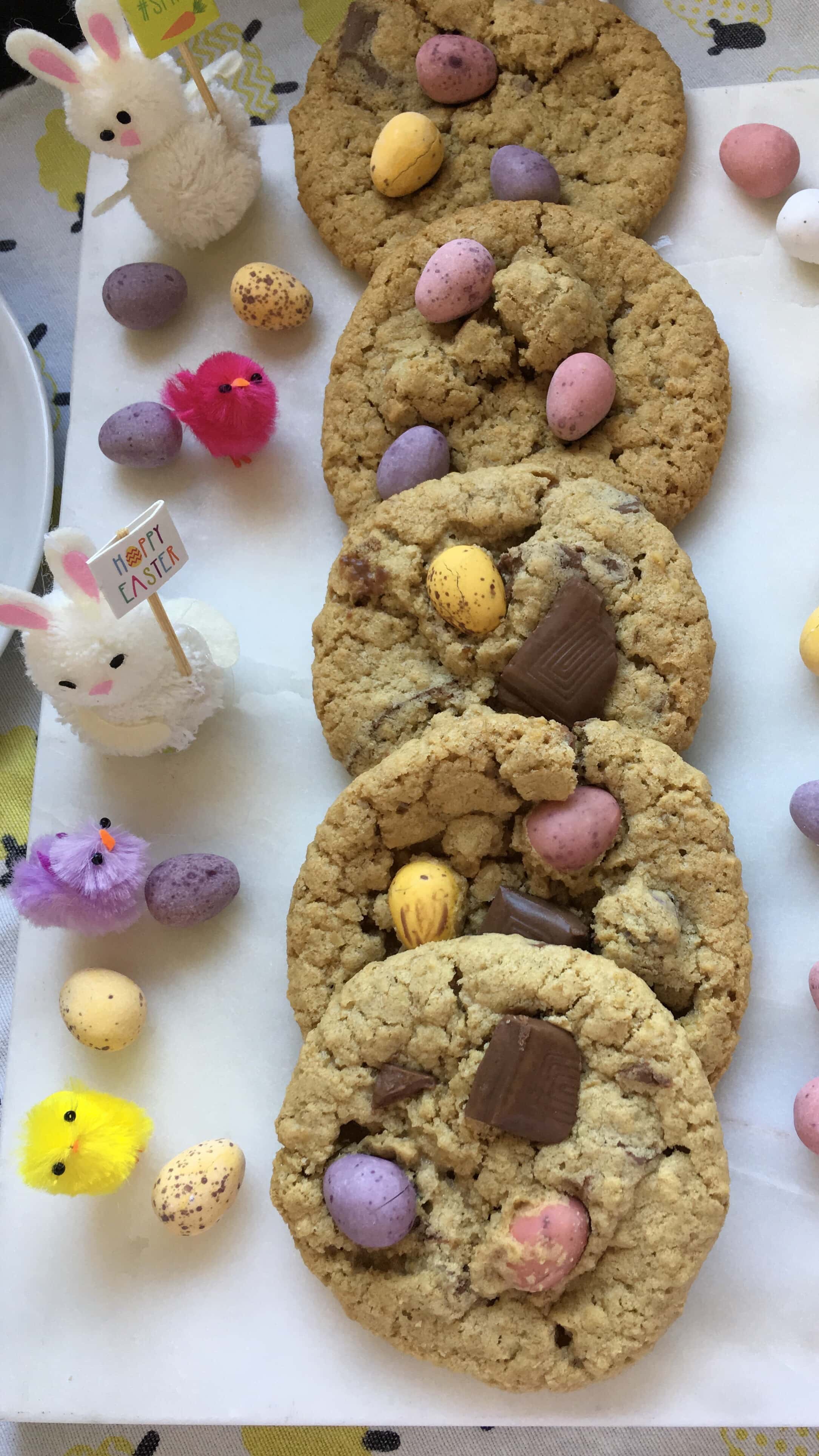 a row of five cookies topped with mini Easter chocolate eggs and decorative Easter chicks and bunnies on a white marbled board.