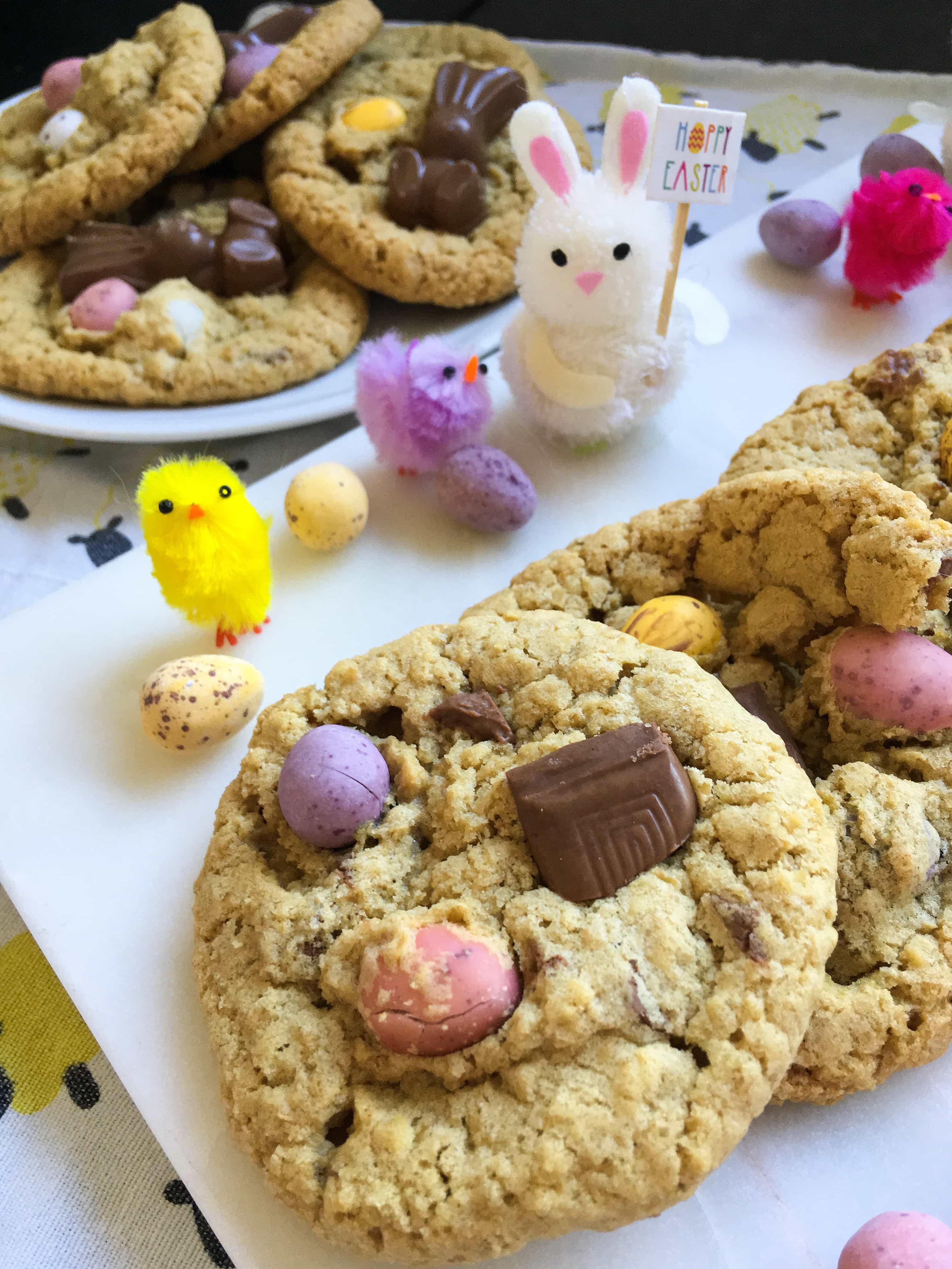 cookies topped with mini coloured chocolate eggs on a white board and decorative Easter chicks and bunny toys. A plate of cookies can be seen in the background.