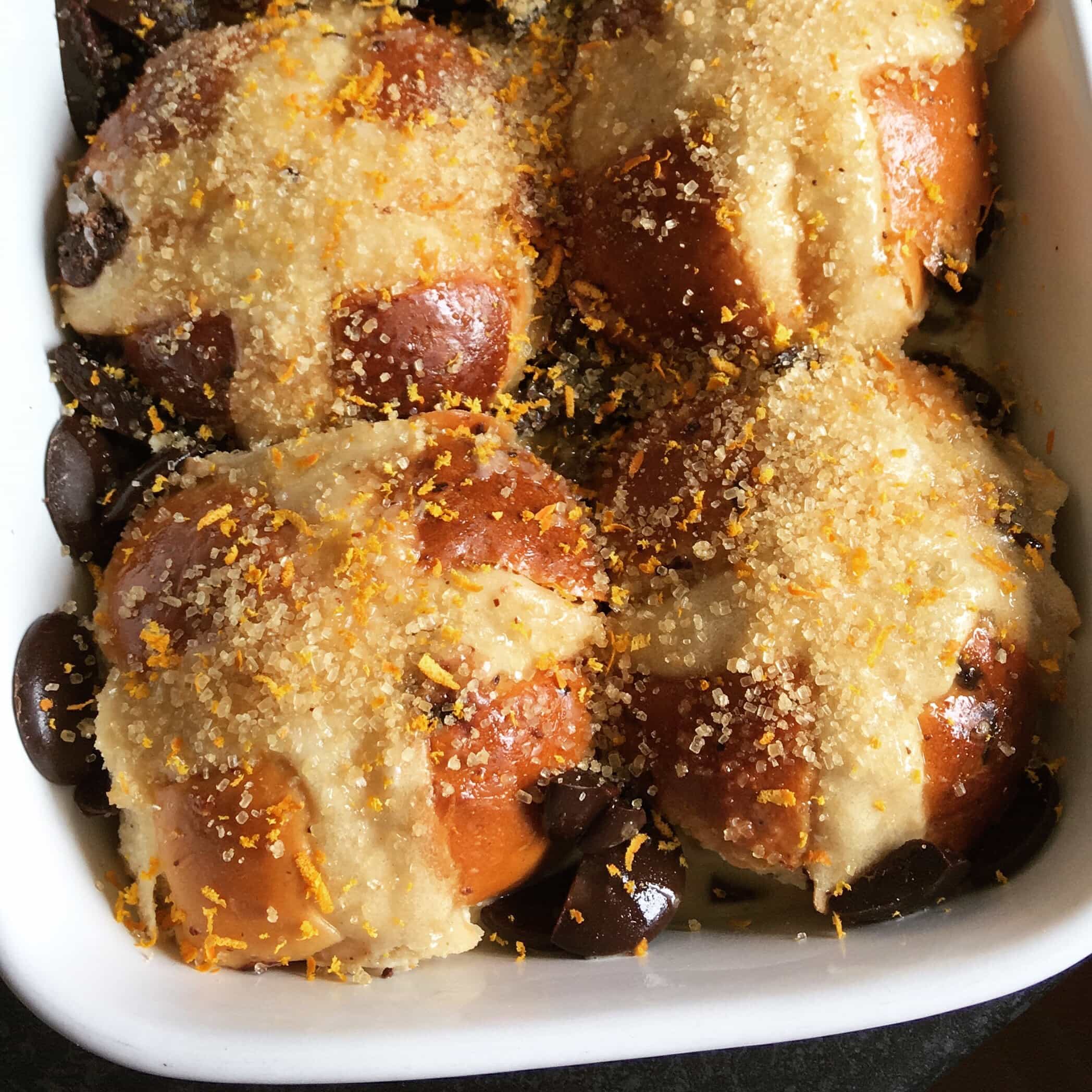 a Hot cross bun pudding in a white dish with melted chocolate, demerara sugar and orange zest.