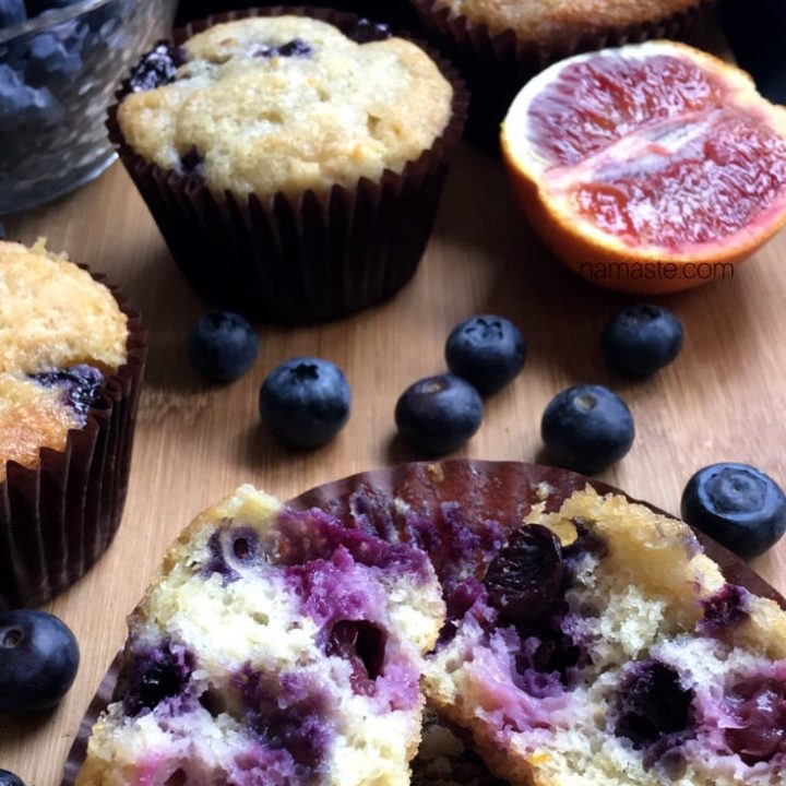 Blueberry muffins on a wooden board. A muffin split in half to display the juicy blueberries sits at the front.
