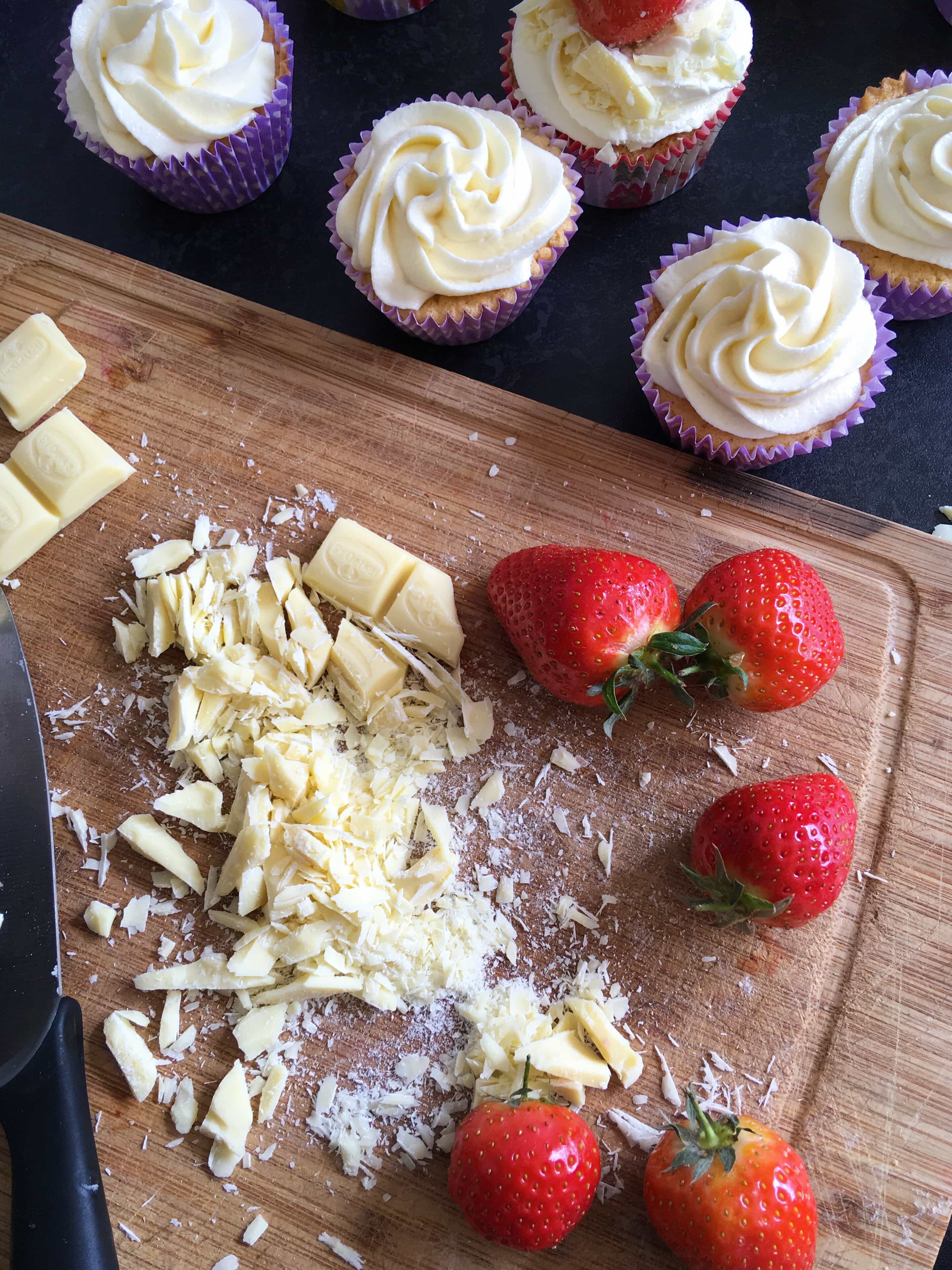 cupcakes topped with white chocolate buttercream and white chocolate pieces and a wooden chopping board with grated white chocolate and fresh strawberries.