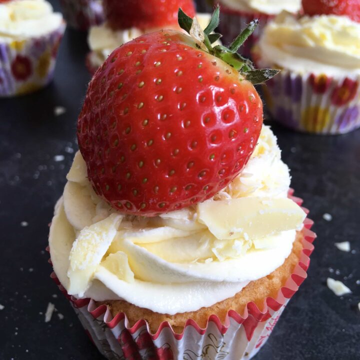 a close up image of a vanilla cupcake topped with a swirl of white chocolate buttercream, white chocolate shards and a fresh strawberry. More cupcakes can be partially seen in the background