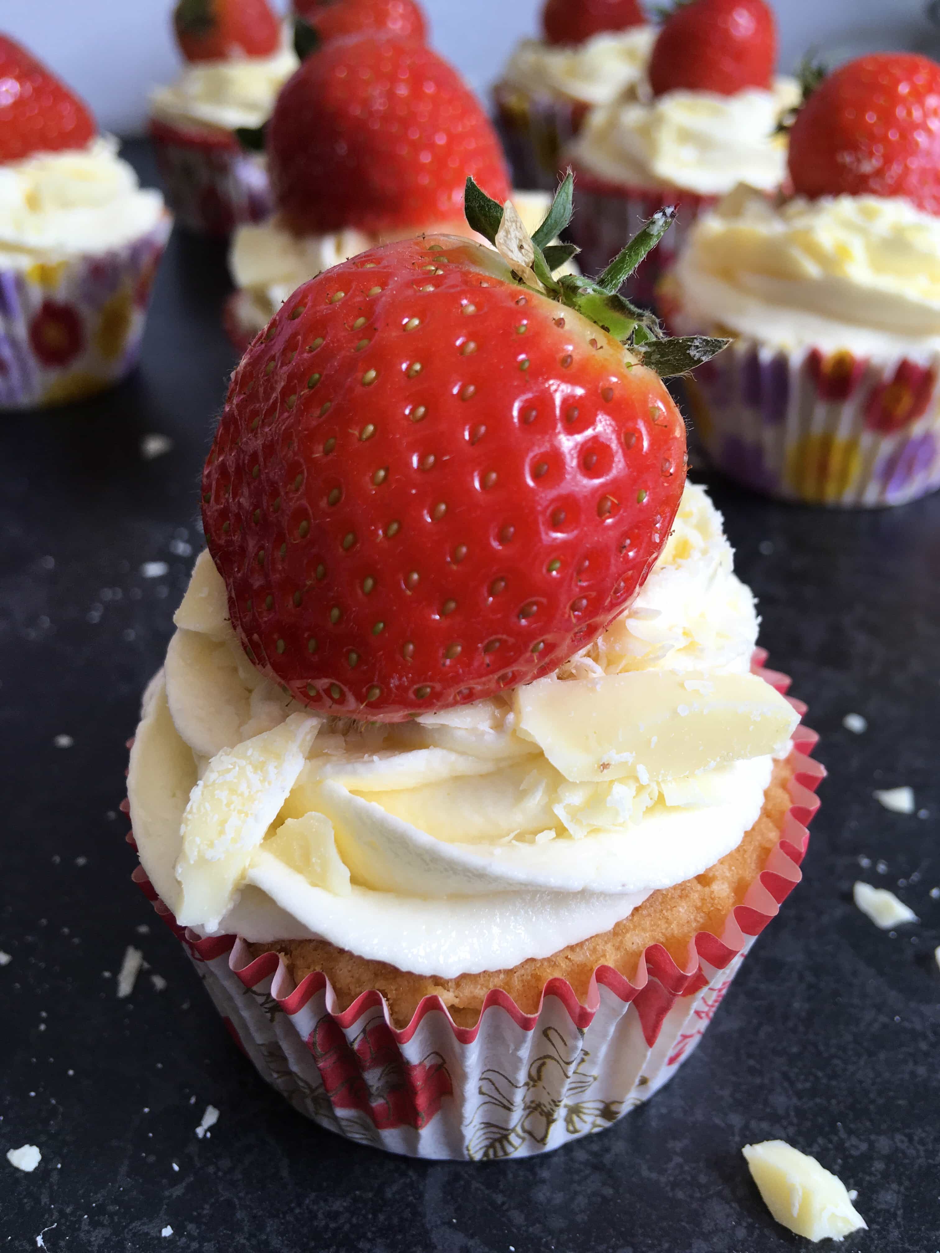 a close up picture of a vanilla cupcake topped with white chocolate frosting, a fresh strawberry and grated white chocolate