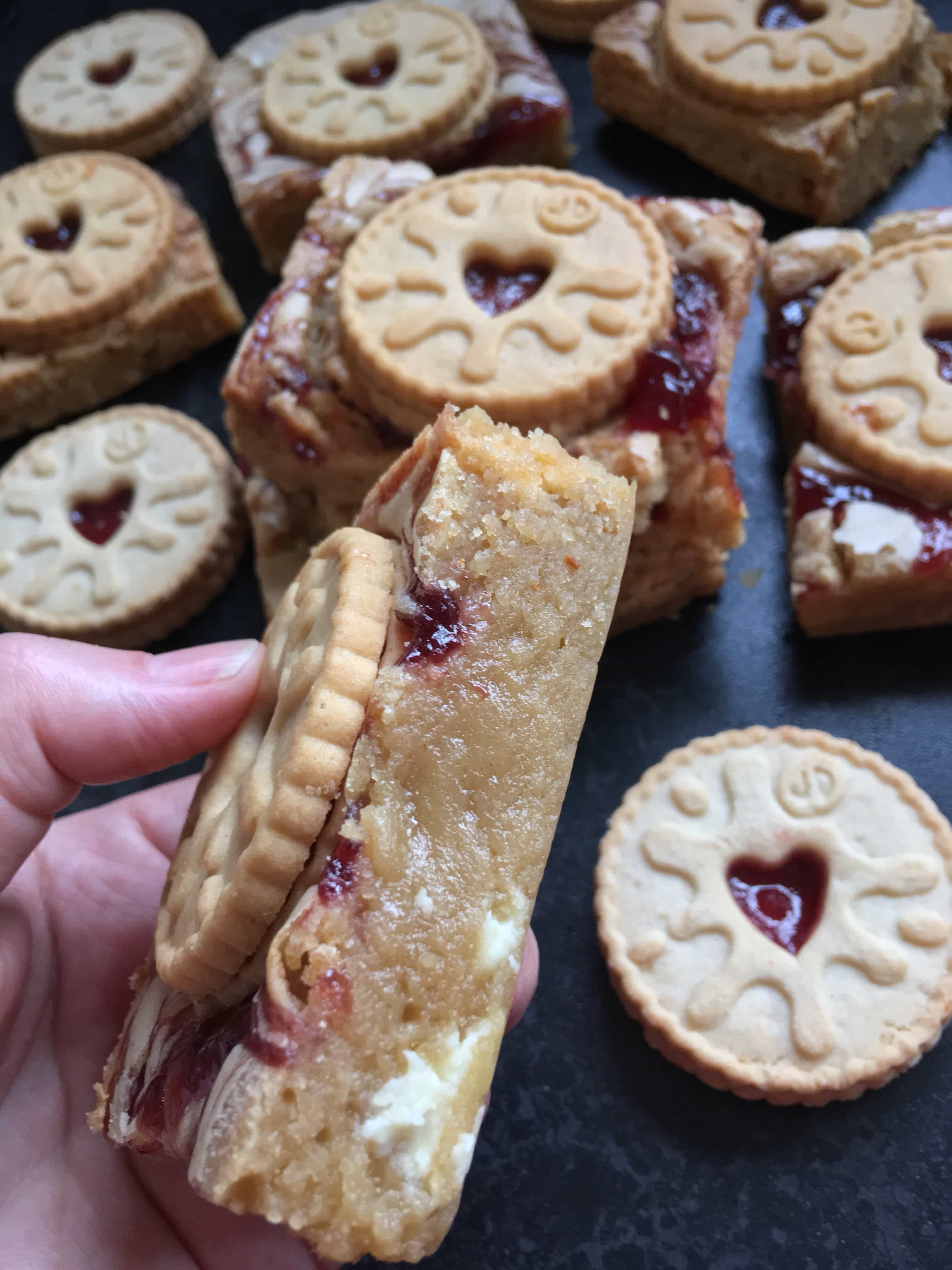 A close up photo of a hand holding a white chocolate blondie with jam swirls and topped with a jammie dodger biscuit