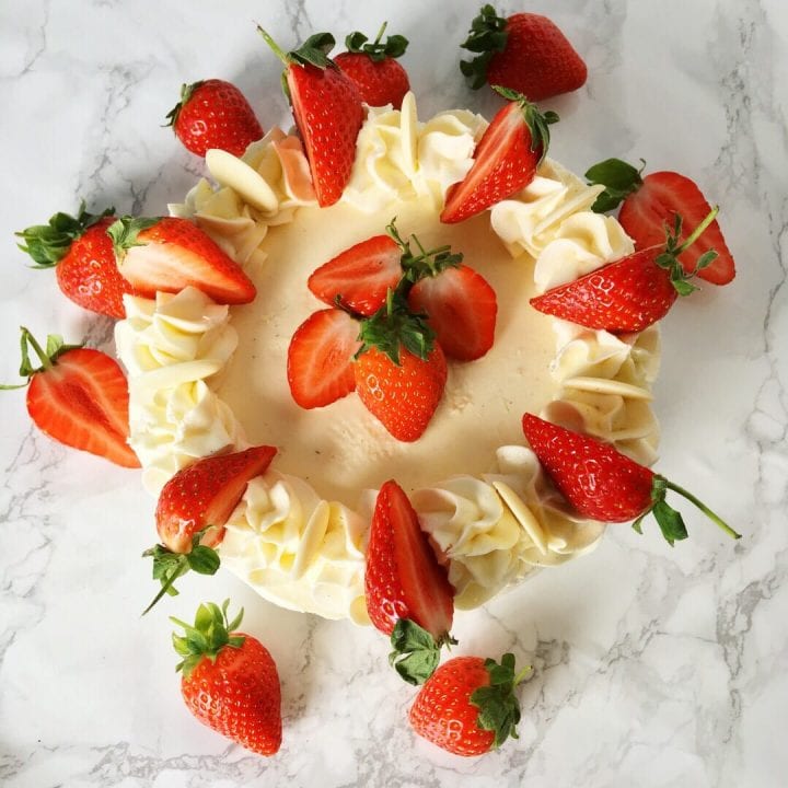 an overhead image of a pretty cake decorated with lemon buttercream frosting, fresh strawberries and white chocolate buttons on a white marbled background.