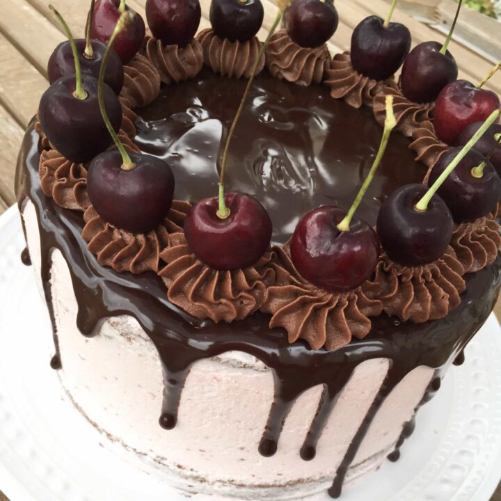 A chocolate cherry drip cake decorated with chocolate buttercream swirls and fresh cherries on a white plate.