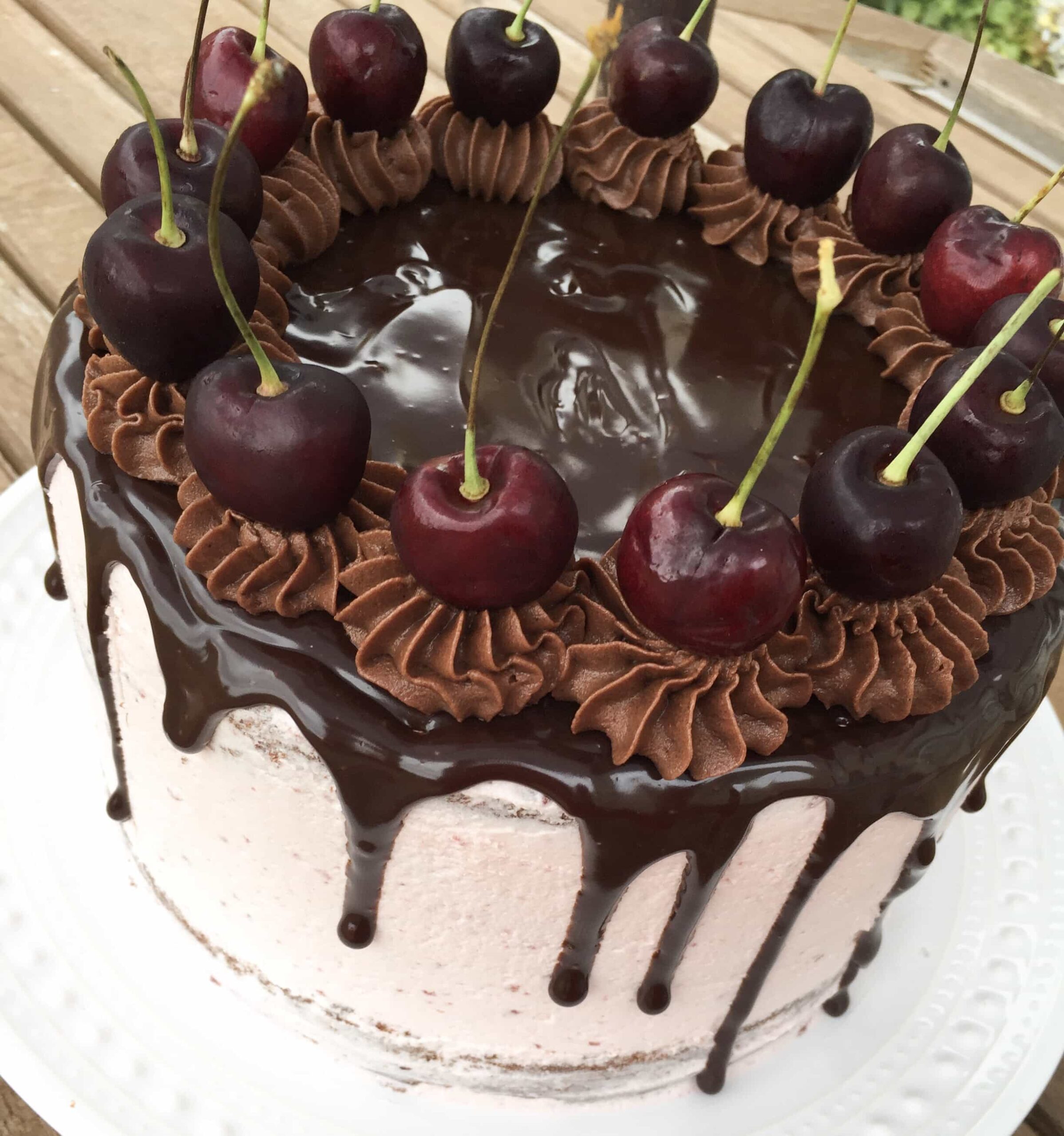 A chocolate cherry drip cake decorated with chocolate buttercream swirls and fresh cherries on a white plate.