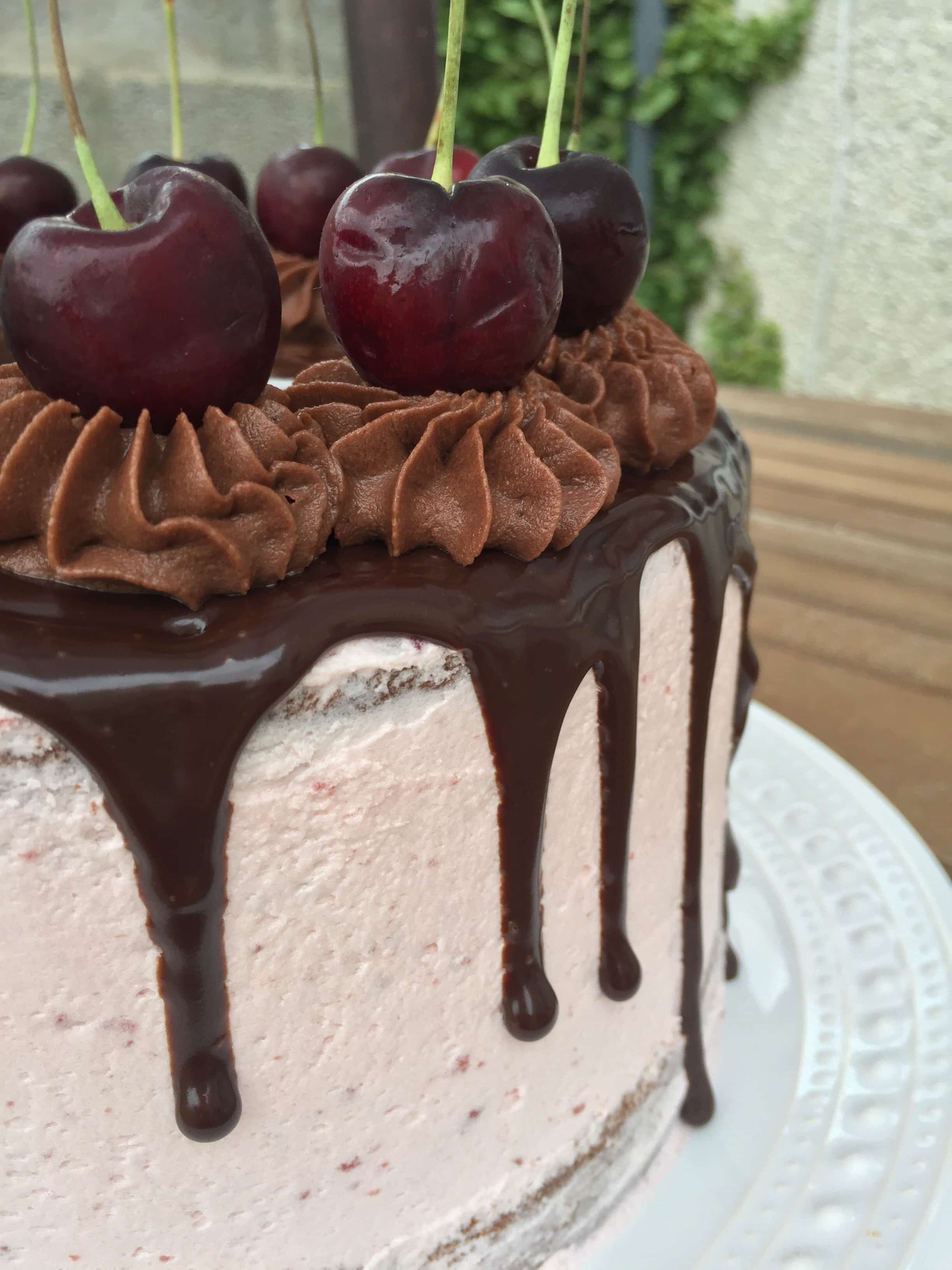 a close up photo of a chocolate cherry cake with a chocolate ganache drip running down the sides and topped with fresh cherries on a wooden table