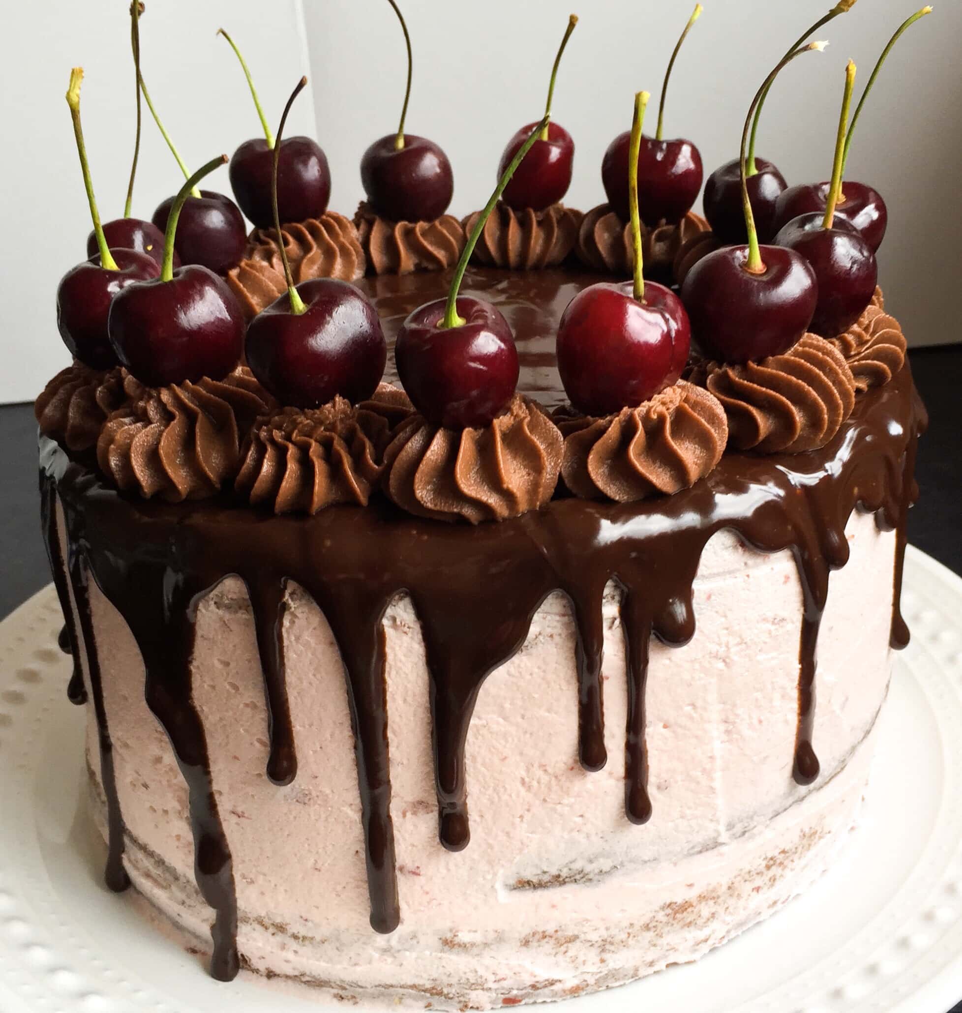 A picture of a chocolate cake decorated with cherry buttercream, a chocolate ganache drip and topped with fresh cherries on a white plate