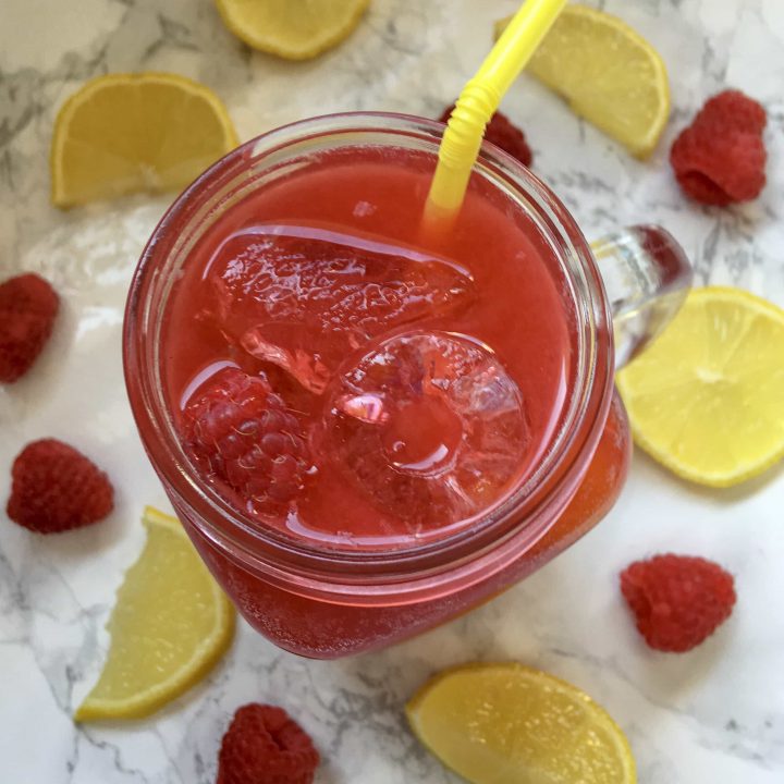 a face down picture of a kilner jug of raspberry Lemonade with ice cubes, fresh raspberries and a yellow straw. Lemon slices and raspberries surround the glass.