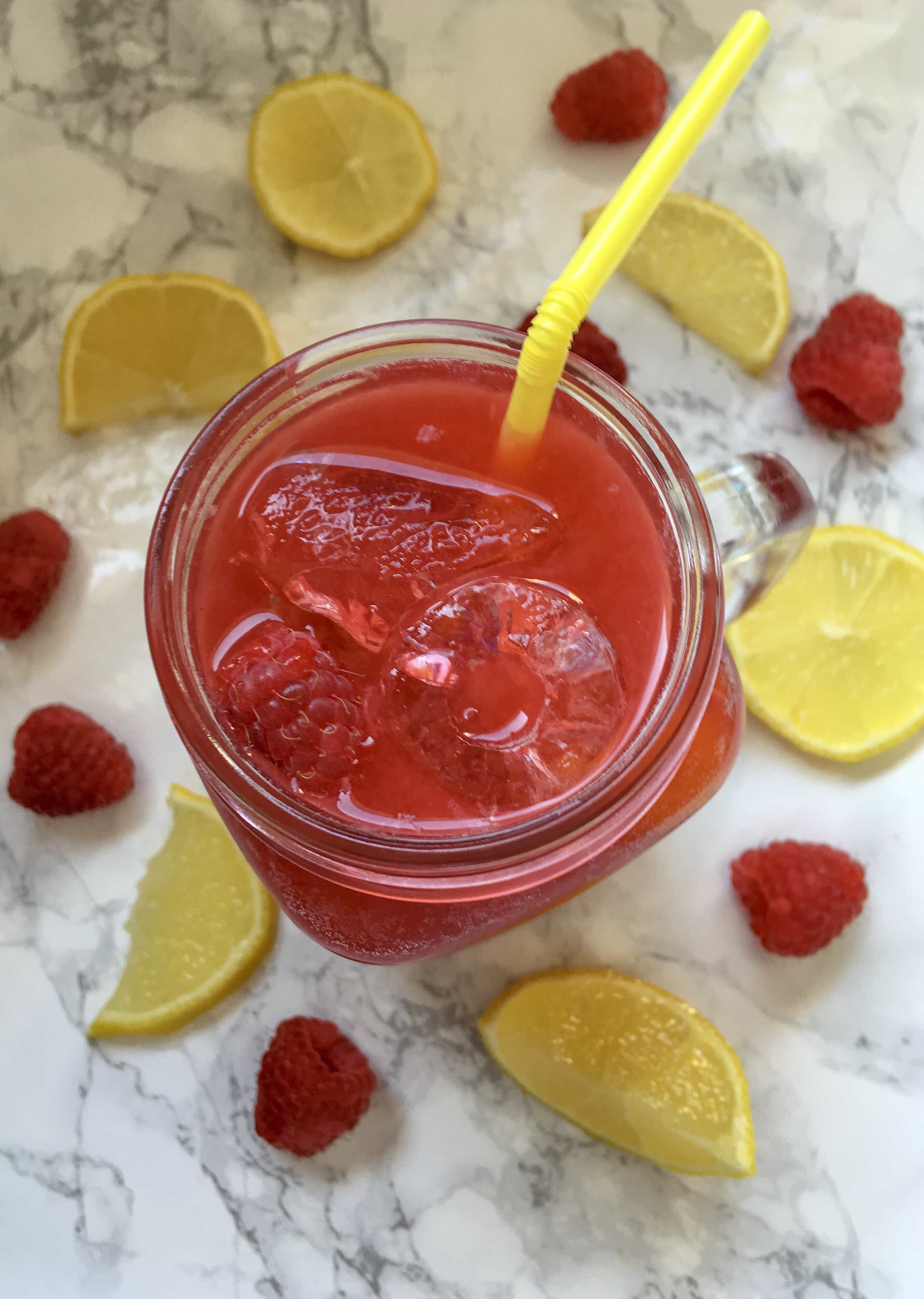 a face down picture of a kilner jug of raspberry Lemonade with ice cubes, fresh raspberries and a yellow straw. Lemon slices and raspberries surround the glass.