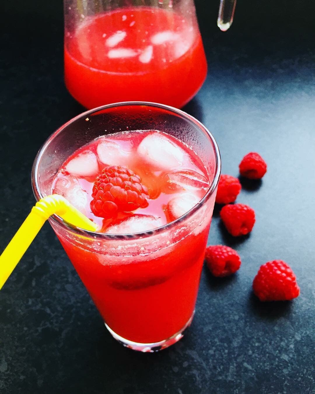 A glass of raspberry lemonade with ice cubes, fresh raspberries and a yellow straw. A jug of the lemonade and fresh raspberries can be seen in the background.