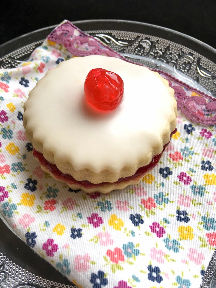 An empire biscuit topped with white icing and a red glace cherry on a mini glass cake stand