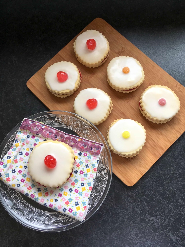 An empire biscuit topped with white icing and a cherry on a glass mini cake stand and six empire biscuits on a wooden board.