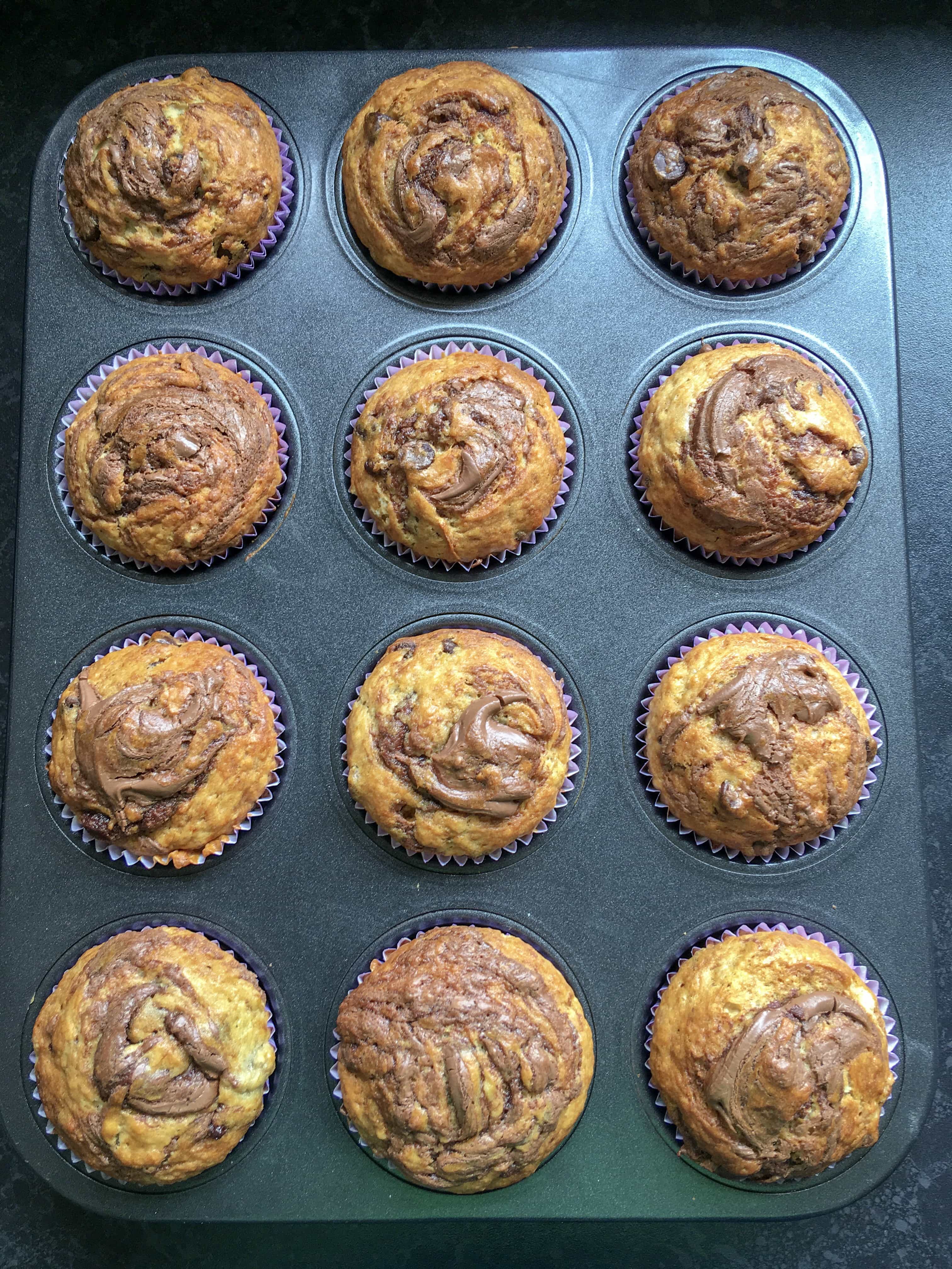 A freshly baked tray of 12 chocolate chip banana muffins with Nutella swirls