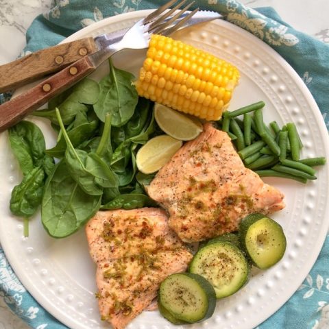 two salmon fillets with a lime and chilli butter with courgettes, spinach and corn on the cob on a white plate/marble background.