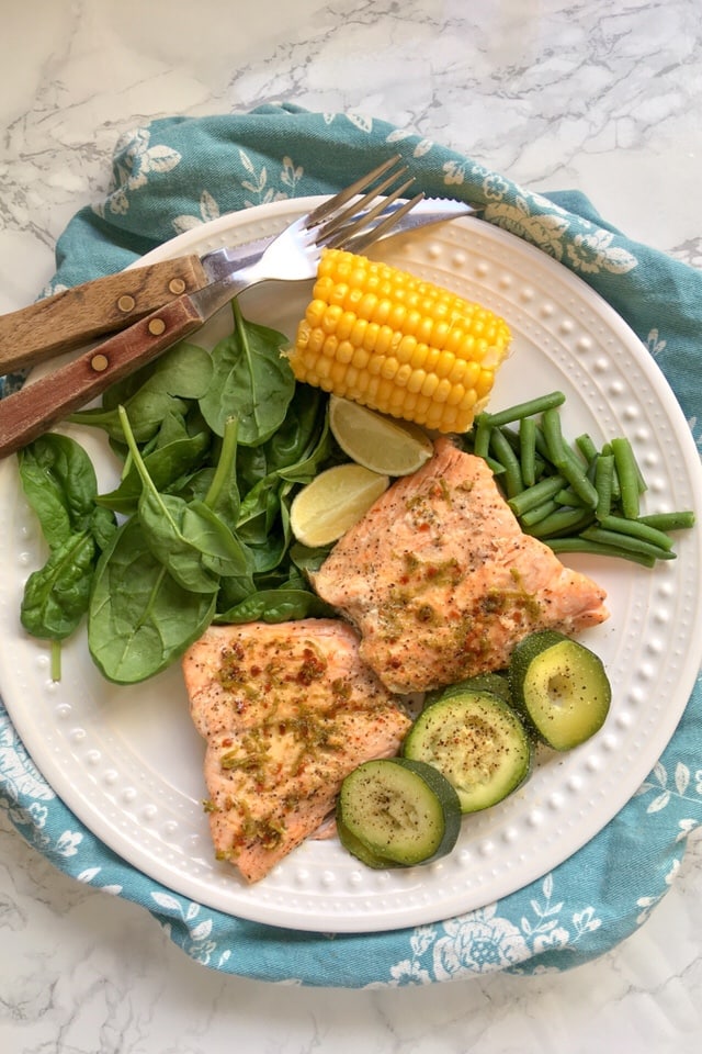 two salmon fillets with a lime and chilli butter with courgettes, spinach and corn on the cob on a white plate/marble background.