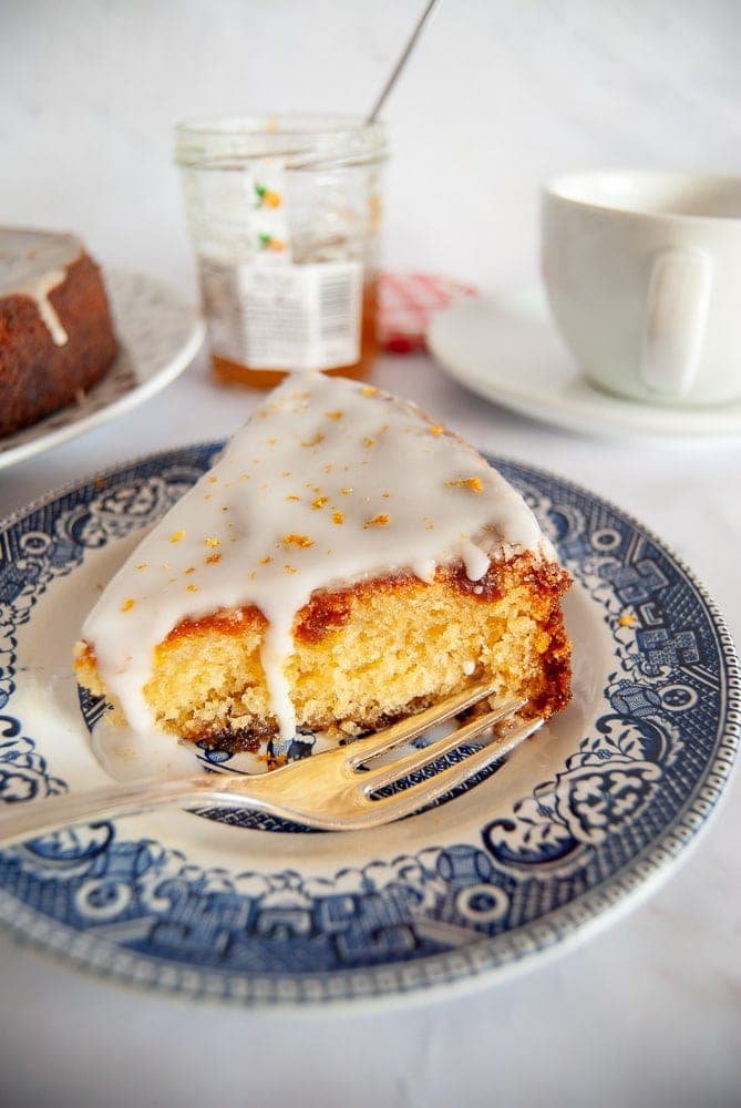 A slice of Orange cake topped with an orange icing glaze and grated orange zest on a Blue Willow Pattern Plate. An open jar of marmalade and a cup of tea can been seen in the background