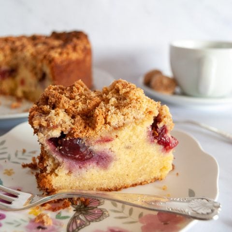 A slice of Plum Amaretti Crumble Cake on a floral plate. The larger cake along with a cup of tea is in the background