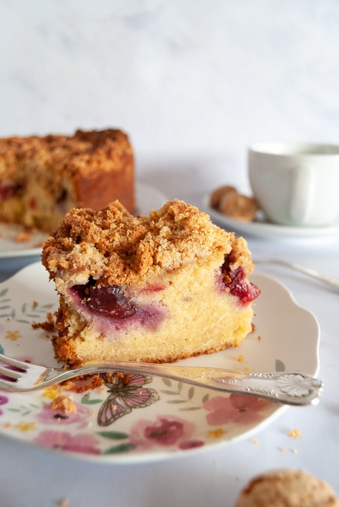 A slice of Plum Amaretti Crumble Cake on a floral plate. The larger cake along with a cup of tea is in the background