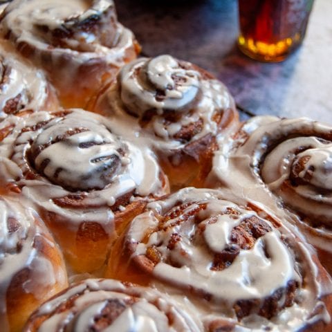 A tray of pumpkin cinnamon rolls on a wire rack/grey background. A black bowl of maple icing and a bottle of maple syrup can be seen in the background.