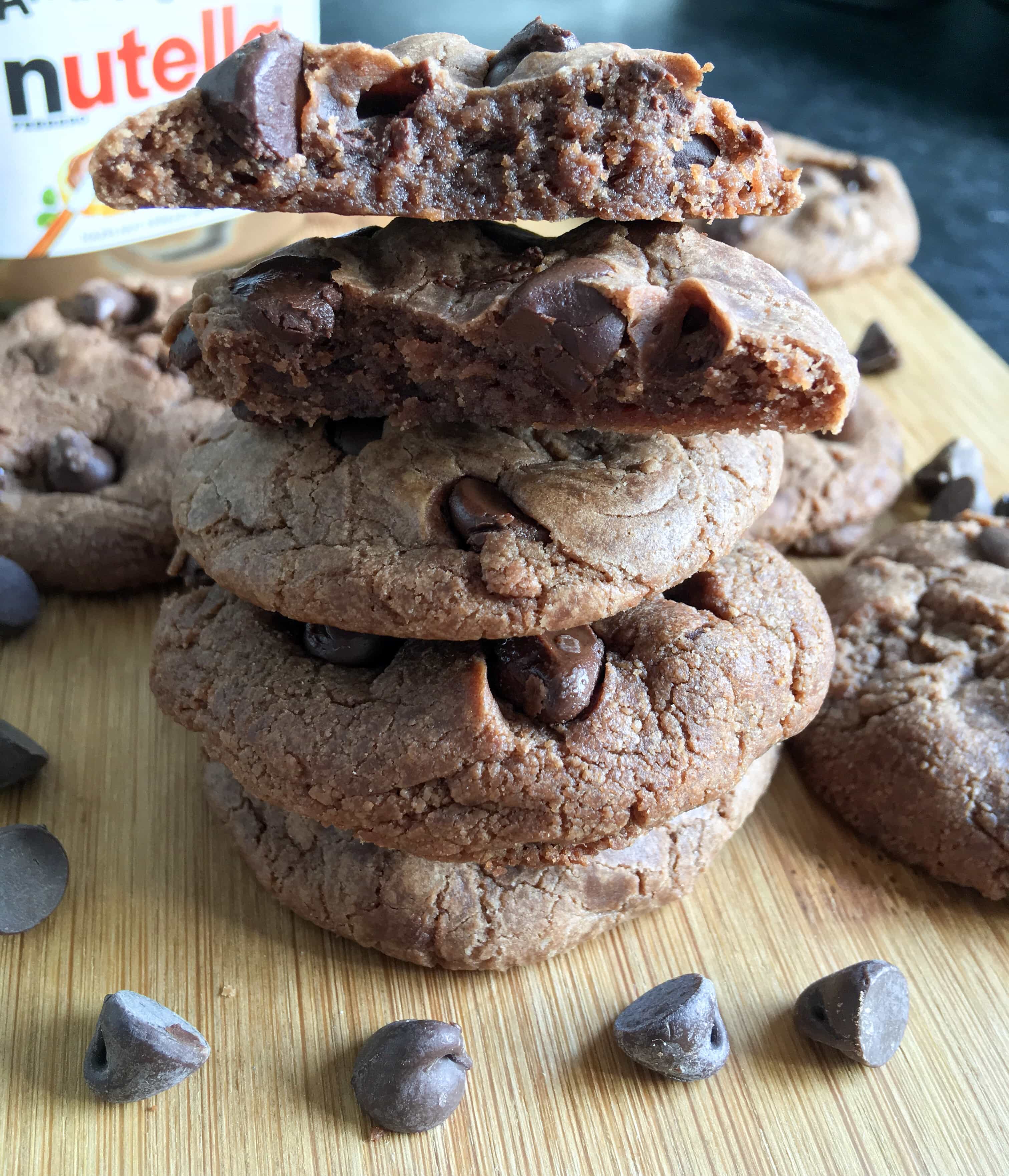 A close up shot of a pile of Nutella cookies with chocolate chips. A jar of Nutella can be partially seen in the background.
