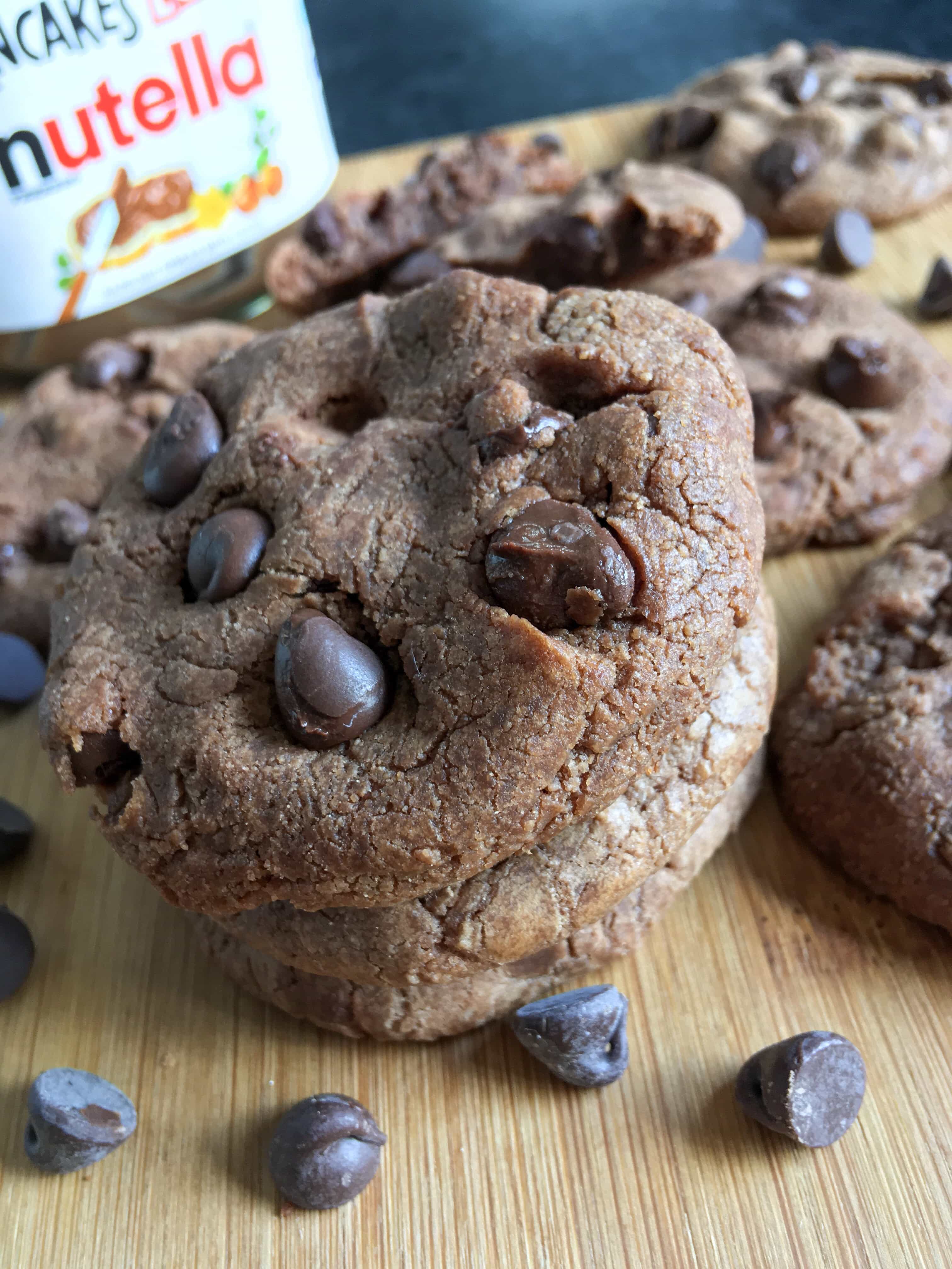 A close up photo of Nutella cookies on a wooden board with dark chocolate chips. A jar of Nutella can just be seen in the background.