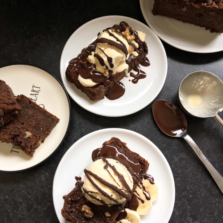A flat lay photo of slices of chocolate banana cake on white plates served with sliced bananas, ice cream and drizzled with chocolate sauce
