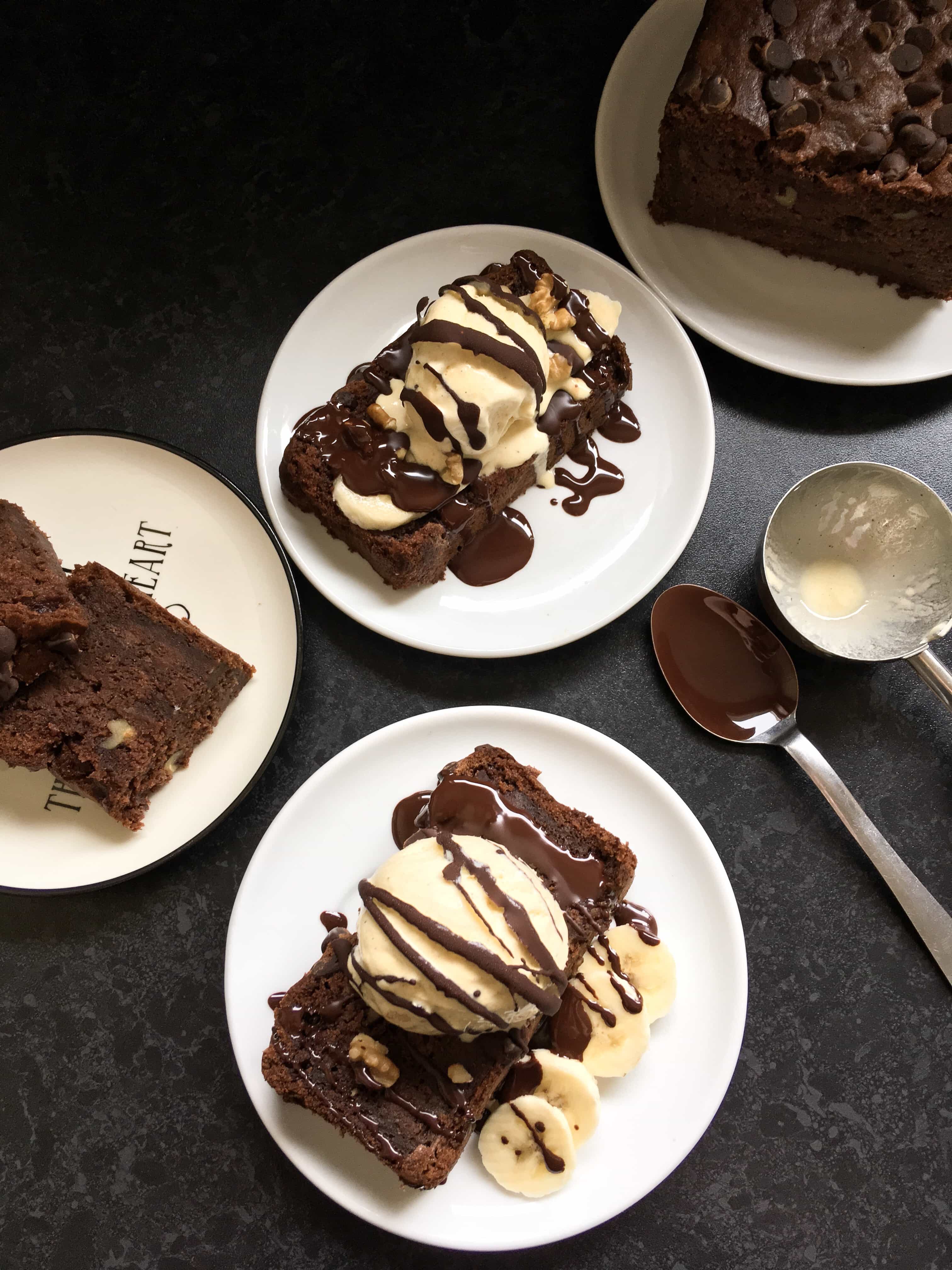 A flat lay photo of slices of chocolate banana cake on white plates served with sliced bananas, ice cream and drizzled with chocolate sauce