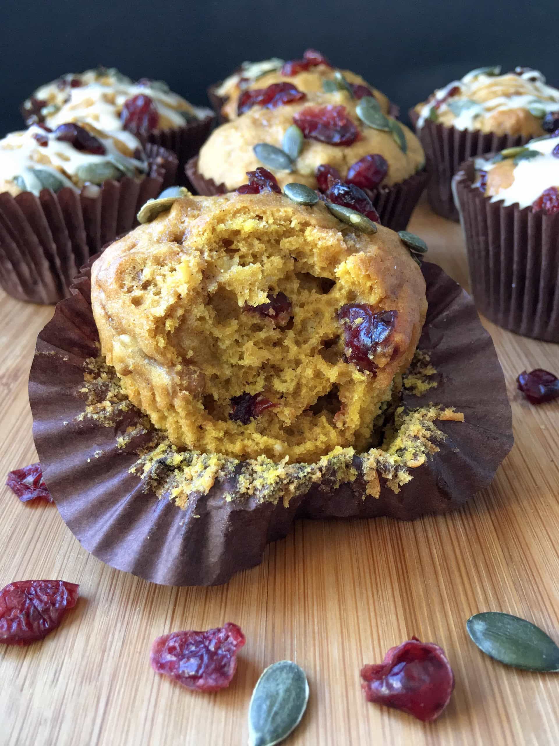 a pumpkin muffin with cranberries on a wooden board, cut open to reveal the inside. Cranberries and pumpkin seeds are scattered around the muffin.