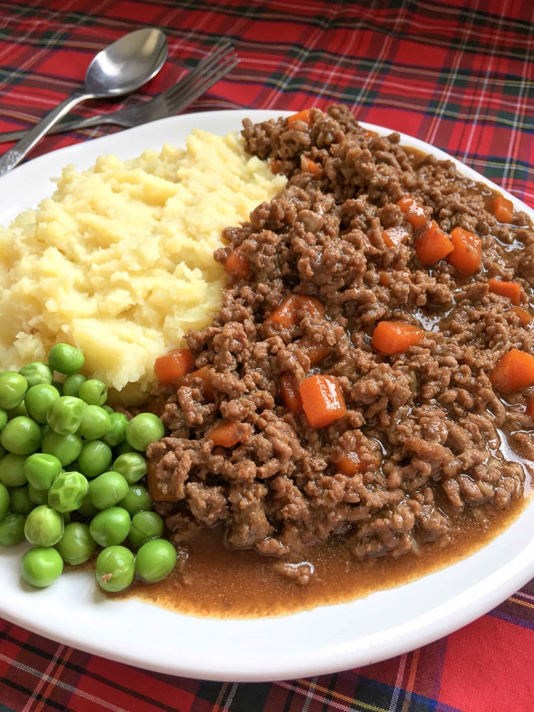 Mince and tatties with peas on a white plate and red tartan background.