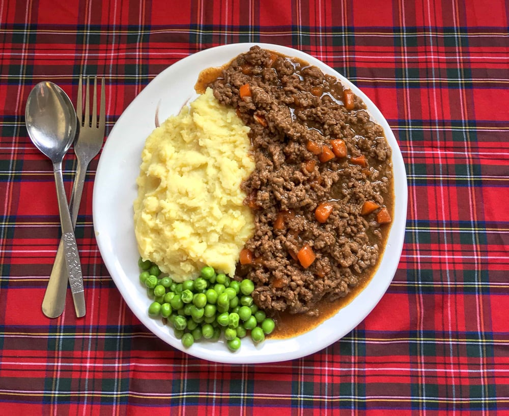 A plate of Scottish mince and tatties with peas on a white plate and tartan background.