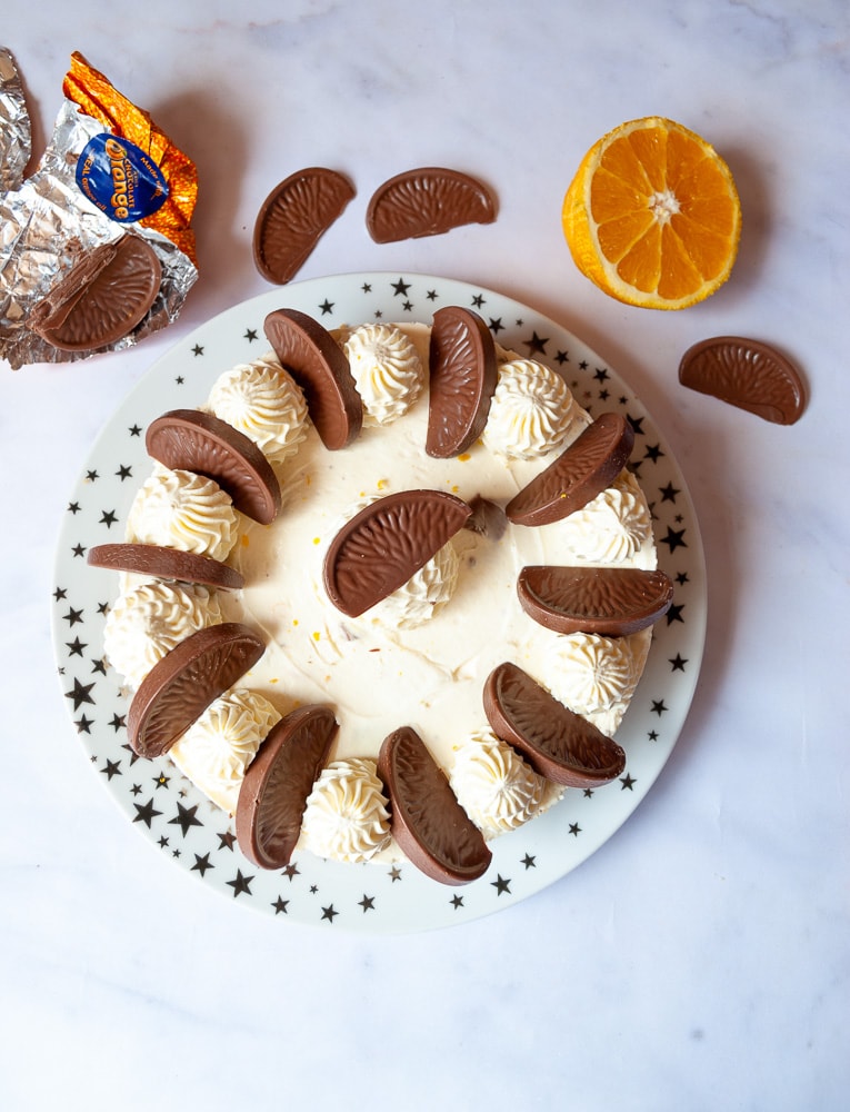 A flat lay photo of a no bake Terry's chocolate orange cheesecake on a gold and white star plate. The cheesecake is decorated with whipped cream swirls and Terry's chocolate orange segments.