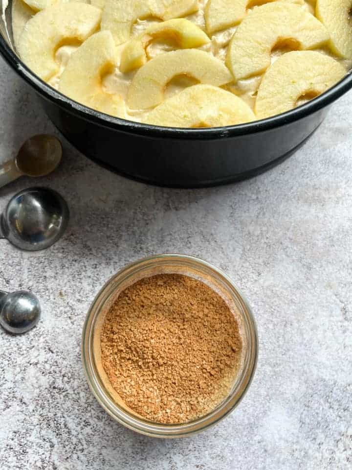 A pot of cinnamon sugar, a set of measuring spoons and an unbaked apple cake topped with apple slices.