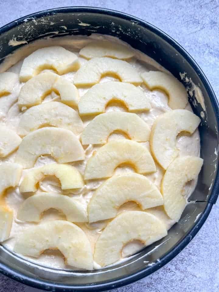 An unbaked apple cake in a black round cake tin topped with apple slices.