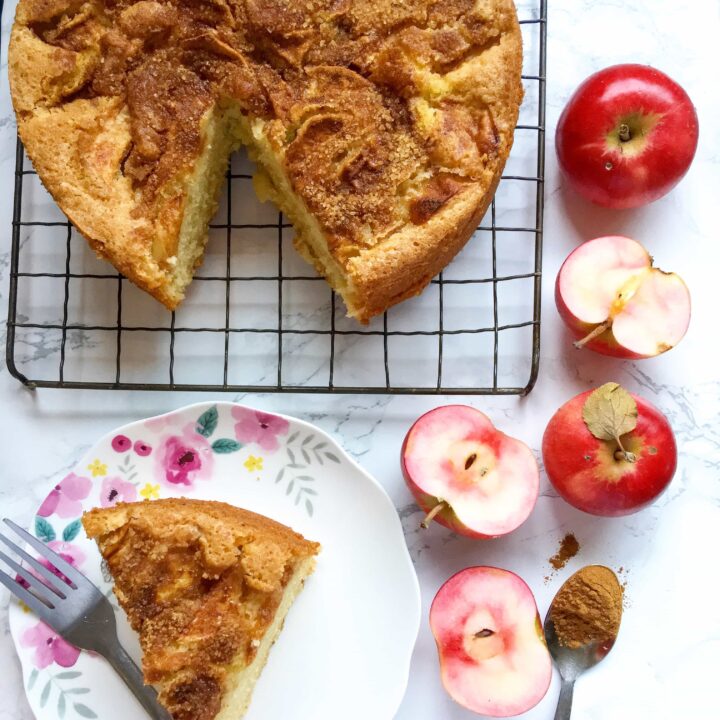 an apple cake topped with crunchy demerara sugar on a silver wire rack and a pretty floral plate with a slice of the cake and a fork. Pink fleshed apples are sitting around the cake along with a spoon of cinnamon.