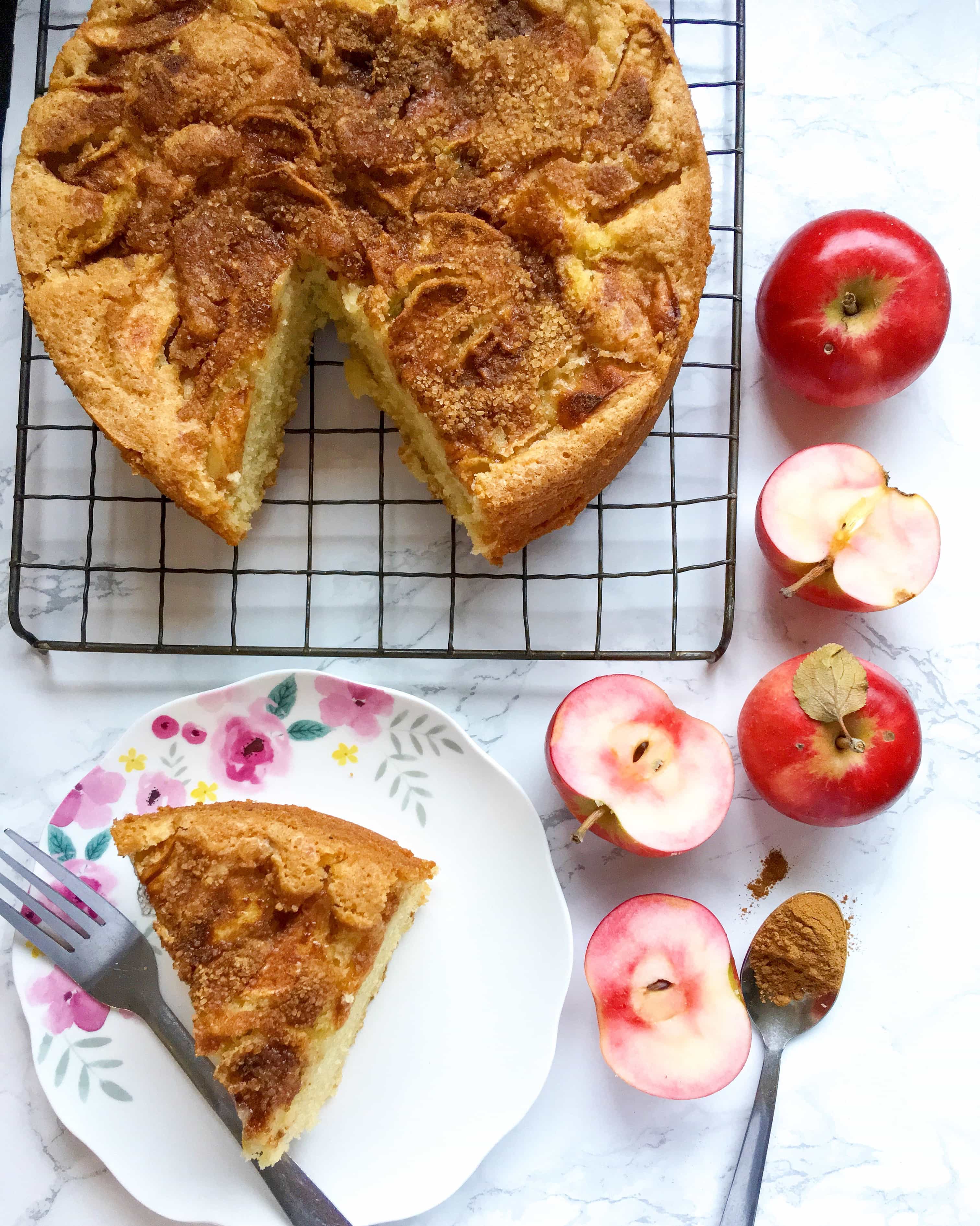 an apple cake topped with crunchy demerara sugar on a silver wire rack and a pretty floral plate with a slice of the cake and a fork. Pink fleshed apples are sitting around the cake along with a spoon of cinnamon.