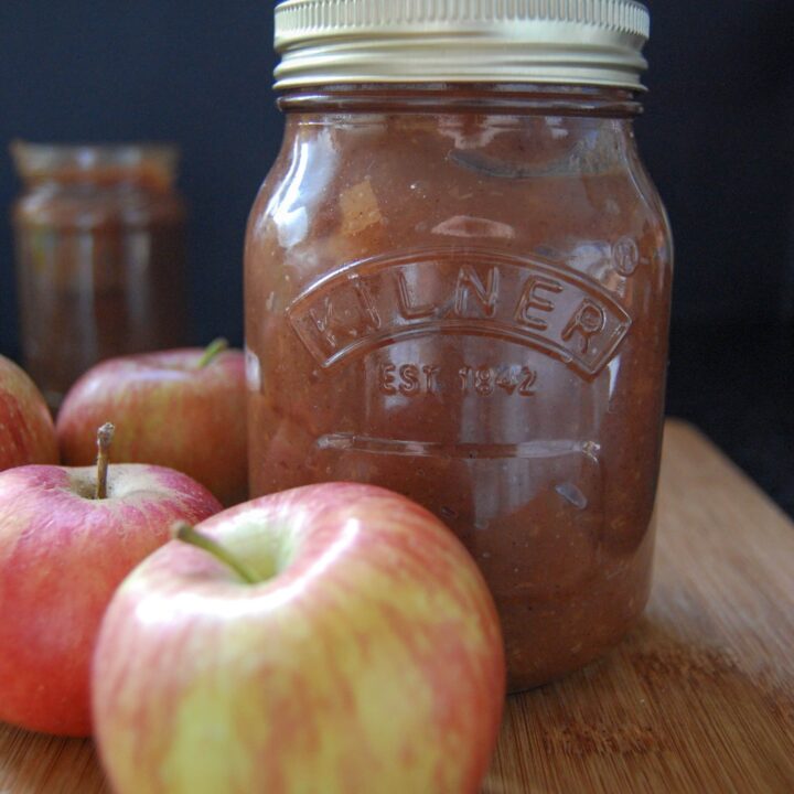 A Kilner jar of apple butter on a wooden board with two red apples,