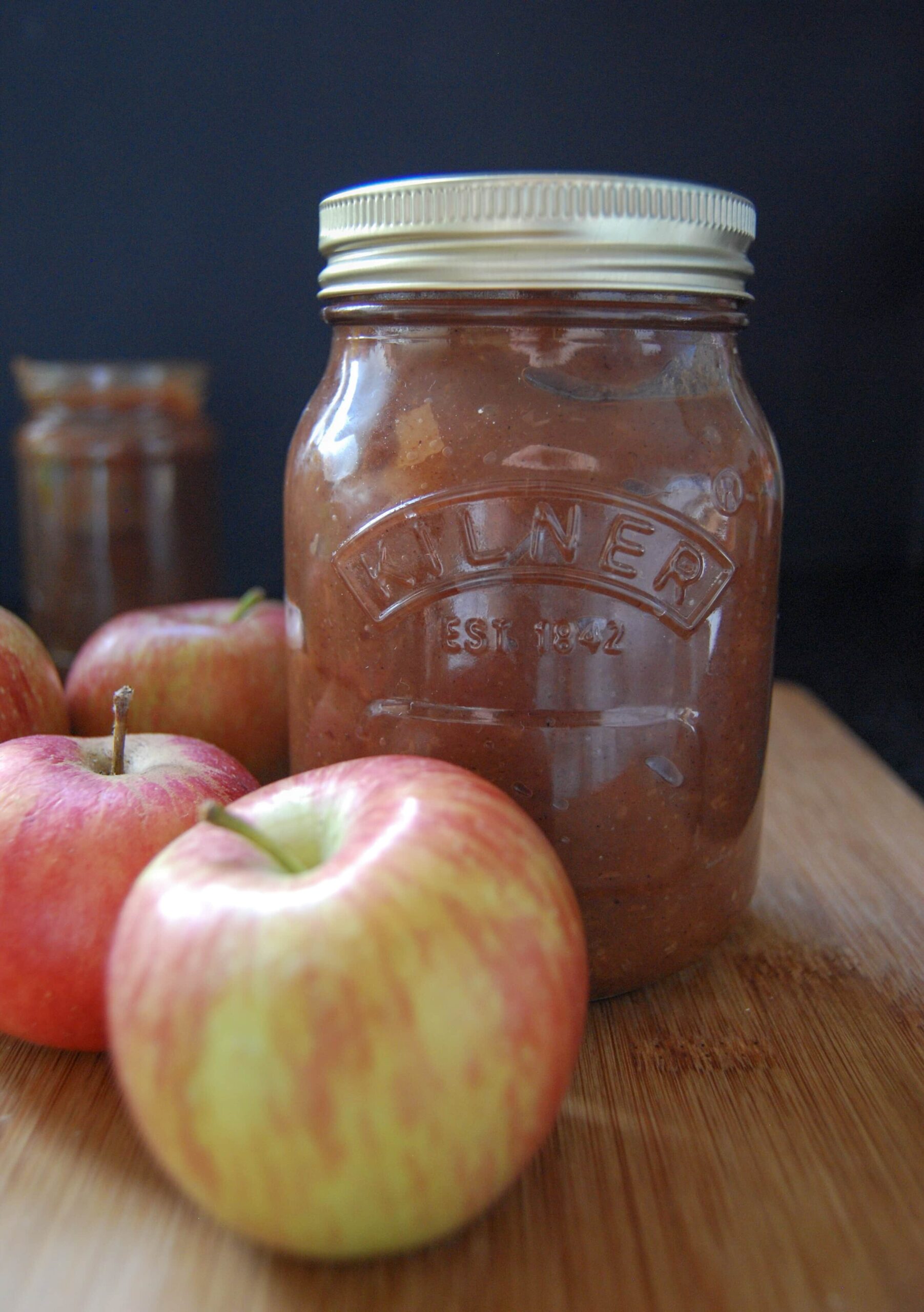 A Kilner jar of apple butter on a wooden board with two red apples,