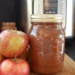 A jar of apple butter and red apples on a wooden board and an Instant Pot in the background