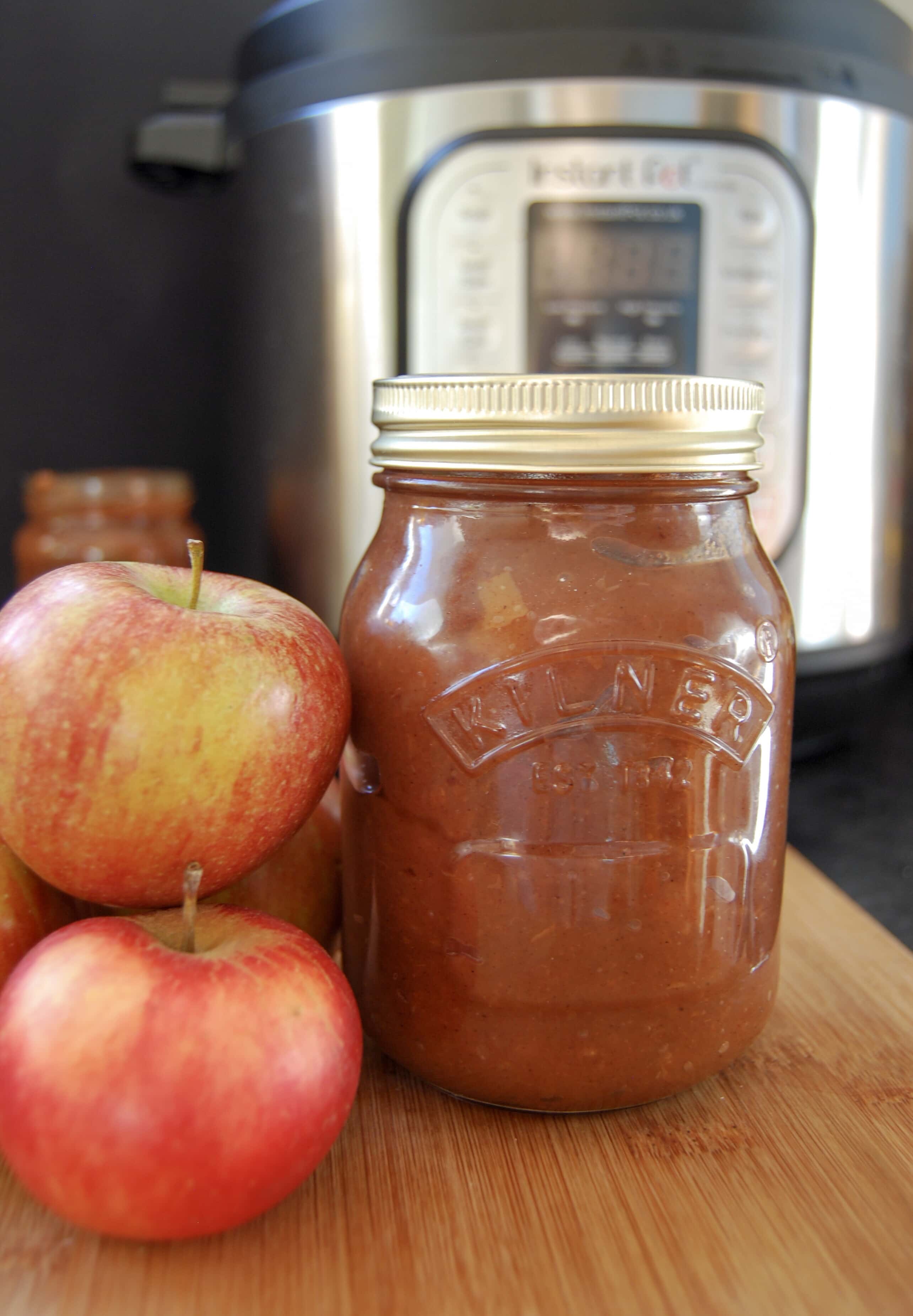 A jar of apple butter and red apples on a wooden board and an Instant Pot in the background