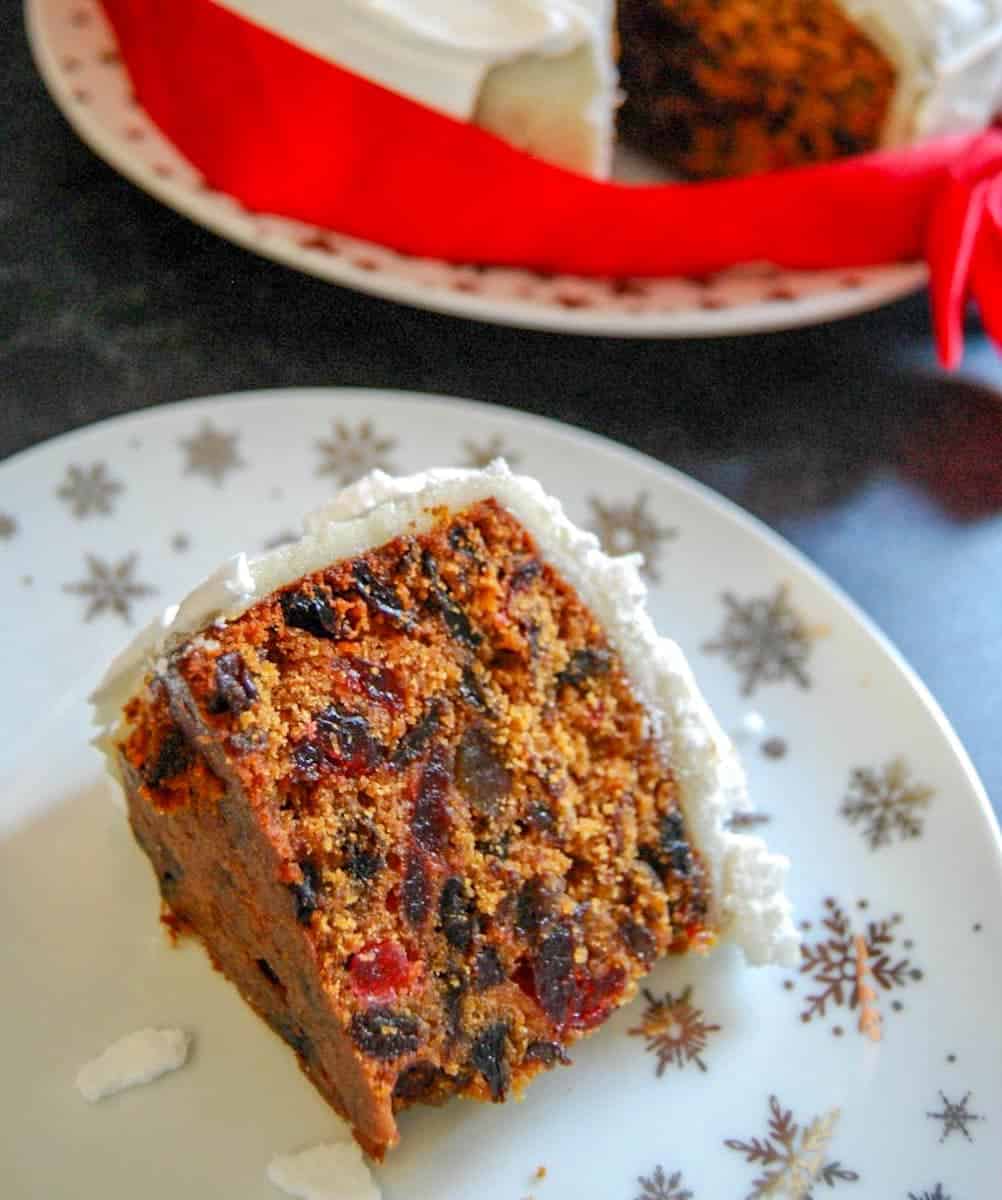 A slice of Christmas cake with marzipan and royal icing on a white and silver snowflake patterned plate. The cake the slice was cut from sits on a white and gold star plate in the background. It has a red ribbon tied around it.