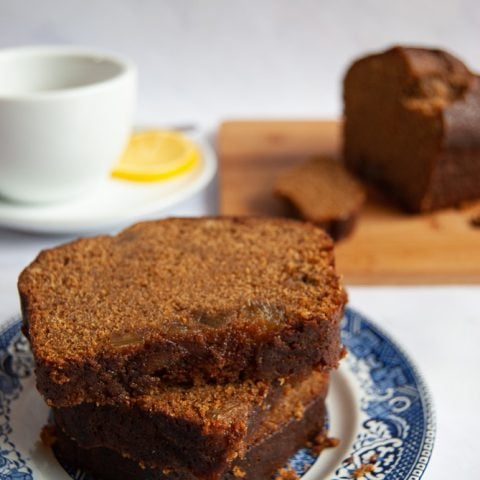Slices of gingerbread loaf cake on a blue willow pattern plate/white background. A cup of tea and the rest of the gingerbread can be seen in the background.